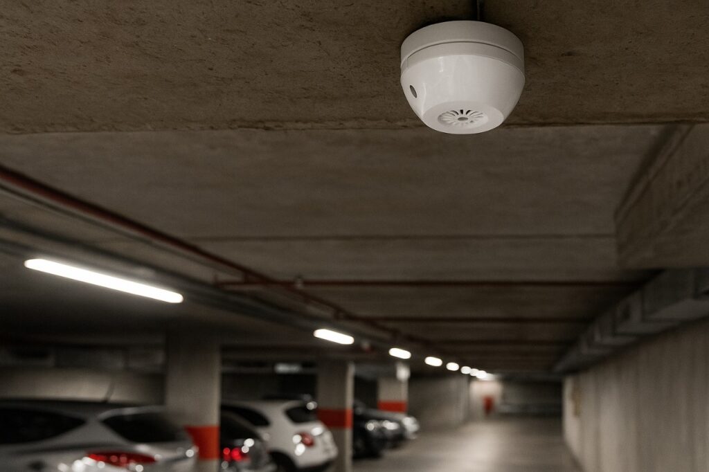 Detector de CO instalado no teto de um estacionamento subterrâneo, com carros estacionados ao fundo e iluminação artificial.