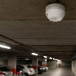 Detector de CO instalado no teto de um estacionamento subterrâneo, com carros estacionados ao fundo e iluminação artificial.