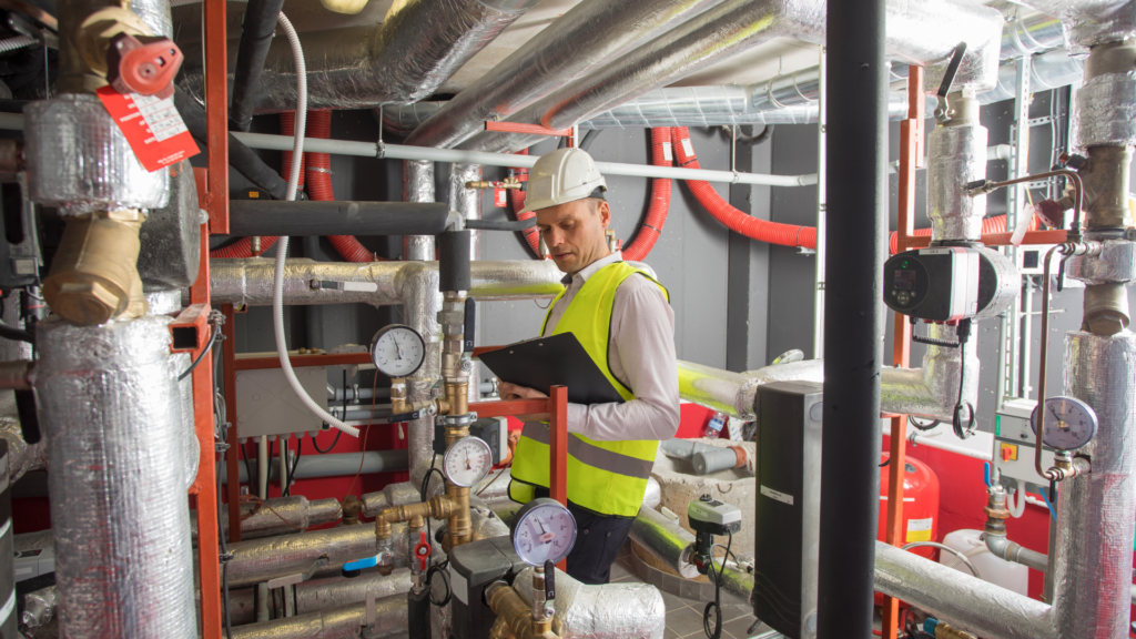 Técnico com colete de segurança e capacete realizando manutenção preventiva em sistema hidráulico e tubulações industriais, utilizando instrumentos de medição e checklist técnico.