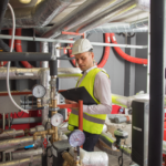 Técnico com colete de segurança e capacete realizando manutenção preventiva em sistema hidráulico e tubulações industriais, utilizando instrumentos de medição e checklist técnico.