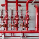 Conjunto de tubulações, válvulas e manômetros pintados de vermelho compondo um sistema de incêndio instalado em ambiente industrial.
