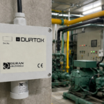 detector de gás fixo instalado em ambiente industrial, ao lado de tubulações de amônia e equipamentos de refrigeração, realizando o monitoramento contínuo para identificar vazamentos de gases e garantir a segurança da operação.